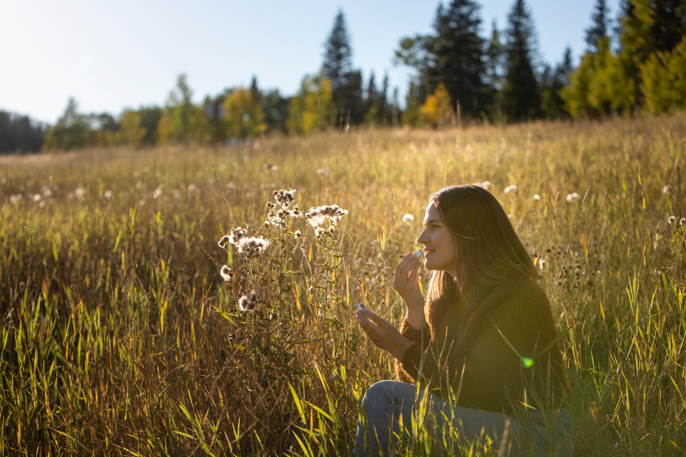 A Canadian woman sitting in a field of tall grass and wildflowers, sourcing all-natural ingredients for lip balm, with trees in the background.