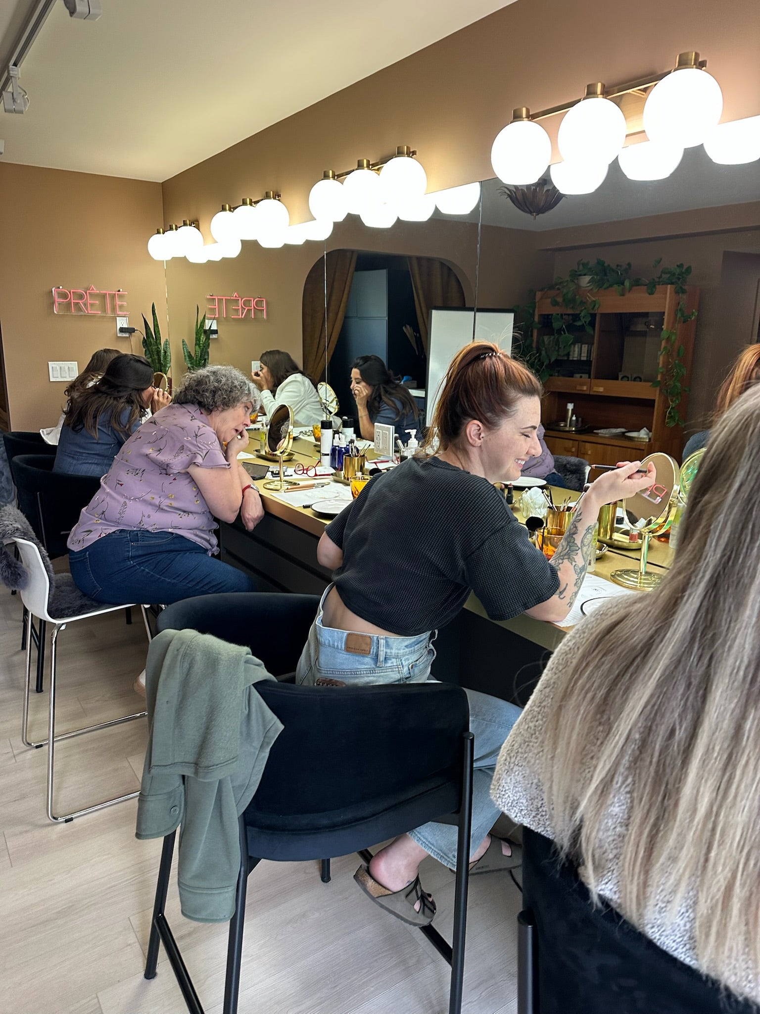 A group of people seated around a vanity table for a makeup workshop in a makeup or beauty salon, with mirrors, bright globe lights, and salon furnishings visible in the room.
